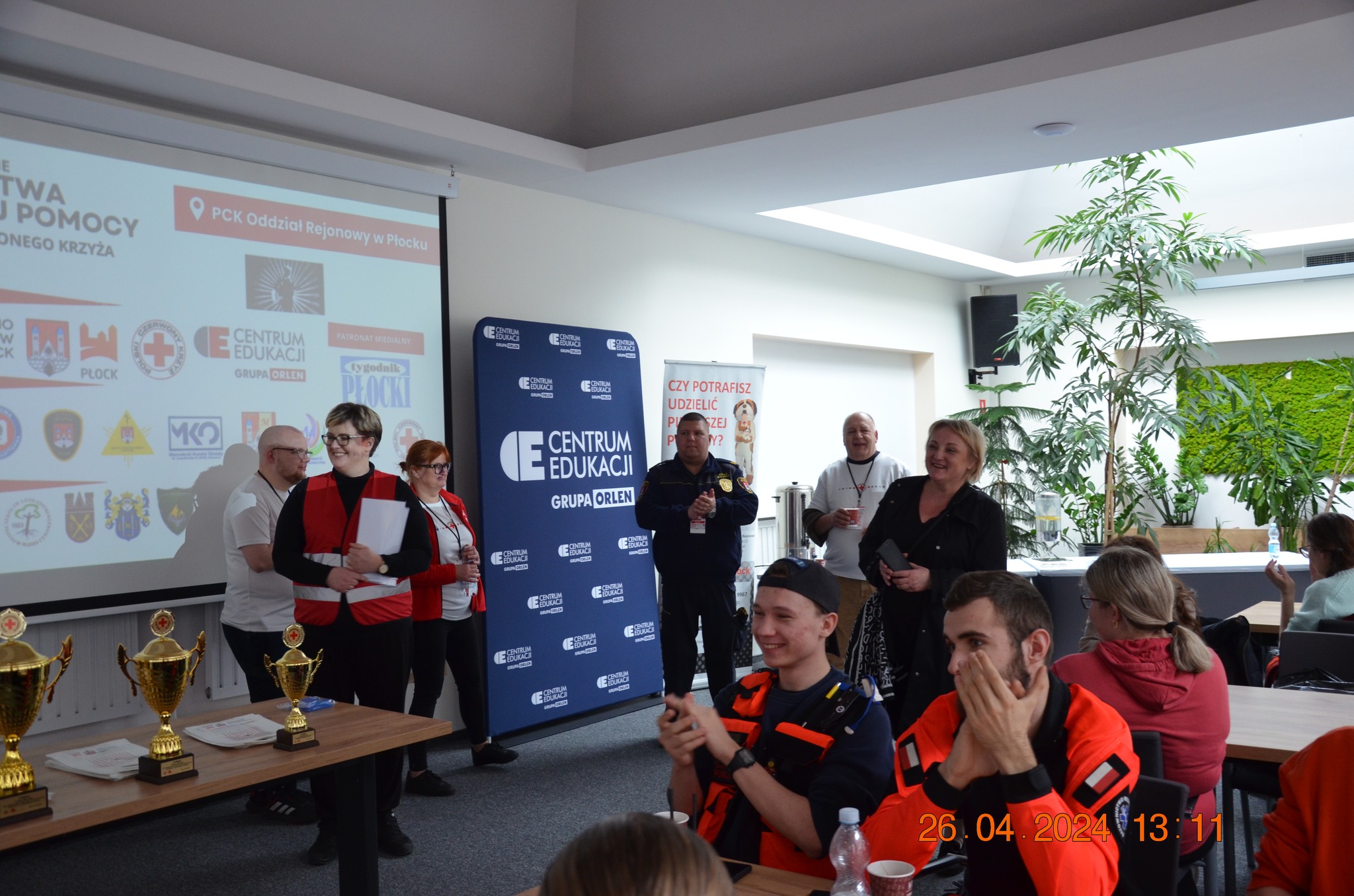 Uczestnicy i organizatorzy etapu rejonowego XXX Ogólnopolskich Mistrzostw Pierwszej Pomocy PCK w Płocku biorą udział w uroczystym podsumowaniu i wręczeniu nagród w sali z prezentacją i pucharami.