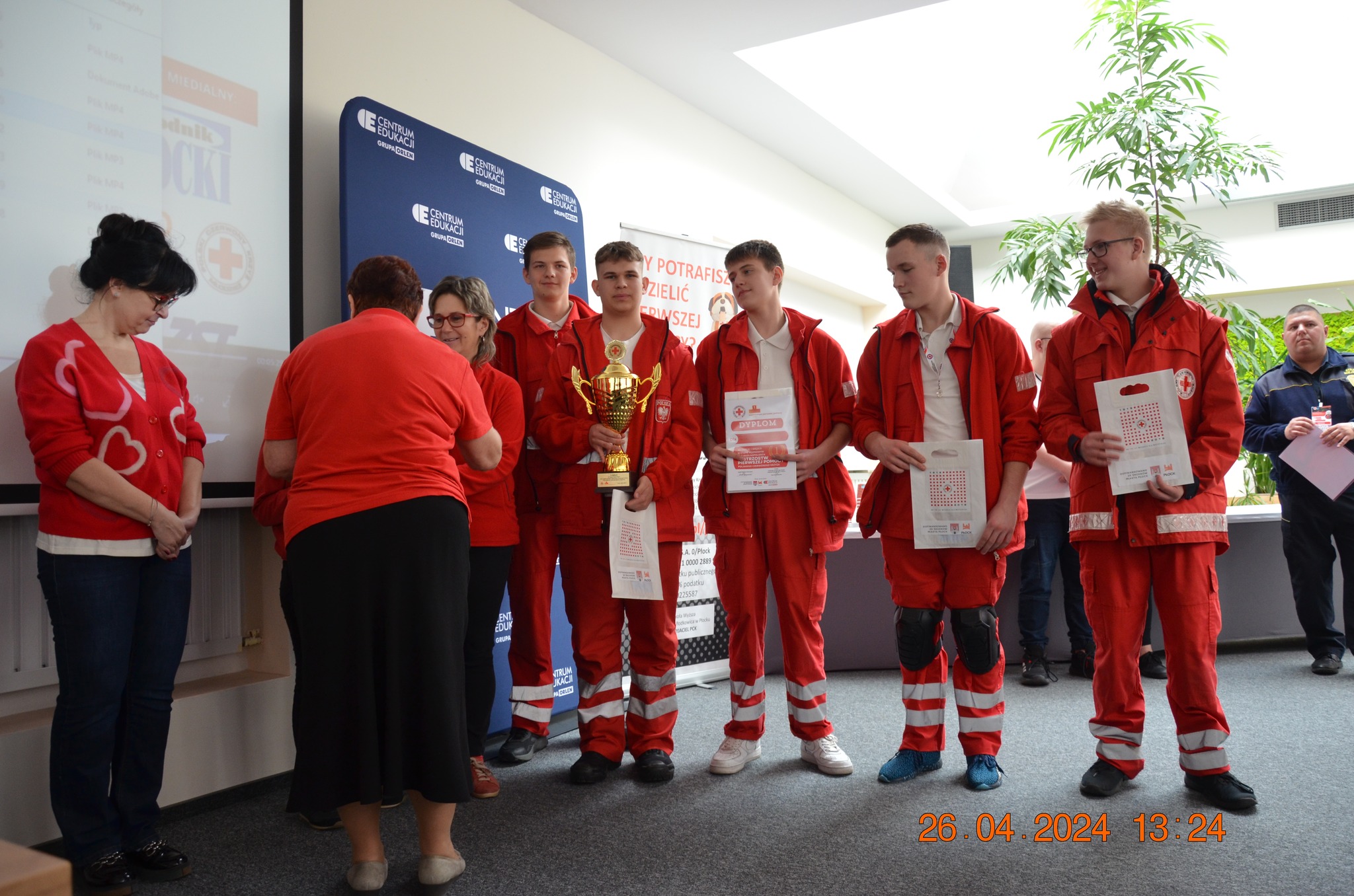 Uczestnicy etapu rejonowego XXX Ogólnopolskich Mistrzostw Pierwszej Pomocy PCK w Płocku stoją w sali podczas ceremonii wręczenia nagród, trzymając puchar, dyplomy i upominki.