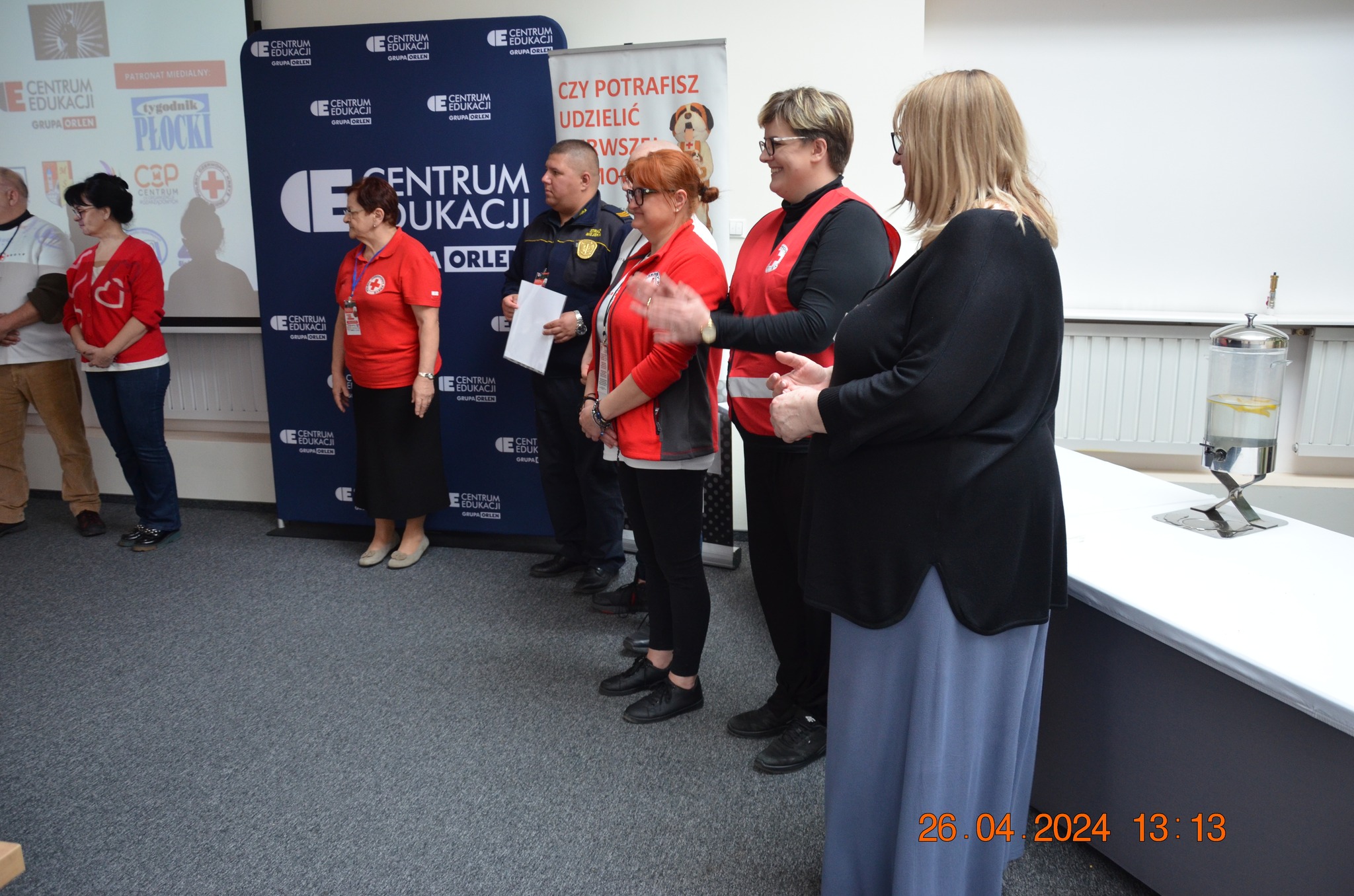 Grupa organizatorów i sędziów XXX Ogólnopolskich Mistrzostw Pierwszej Pomocy PCK w Płocku stoi przed ścianką Centrum Edukacji ORLEN podczas części oficjalnej etapu rejonowego.
