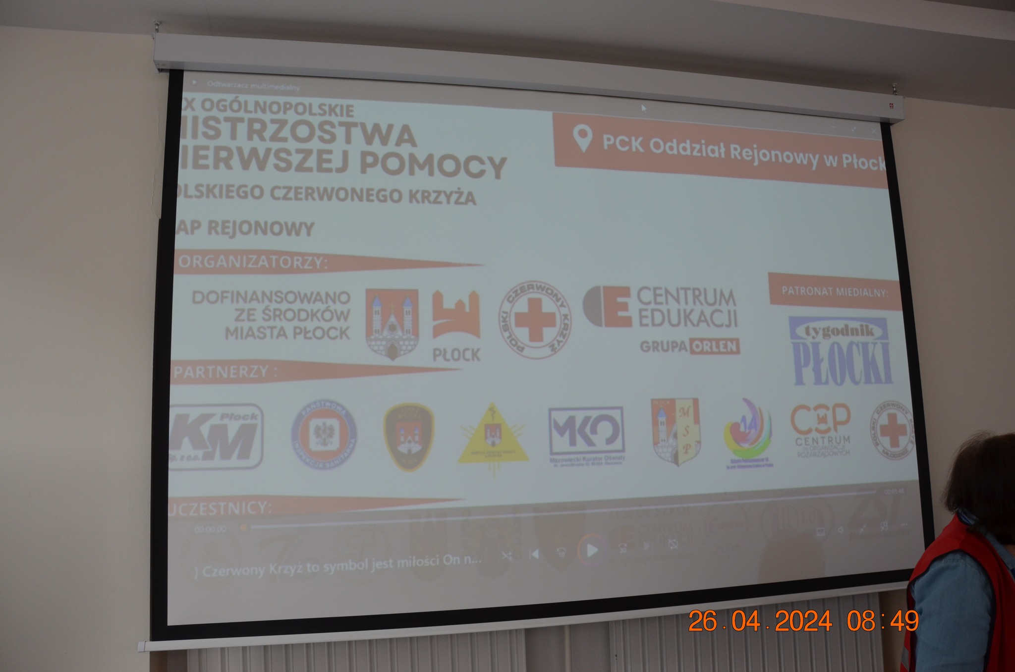 Na zdjęciu widać ekran z prezentacją dotyczącą etapu rejonowego XXX Ogólnopolskich Mistrzostw Pierwszej Pomocy PCK w Płocku, z logotypami organizatorów, partnerów i patronów wydarzenia.