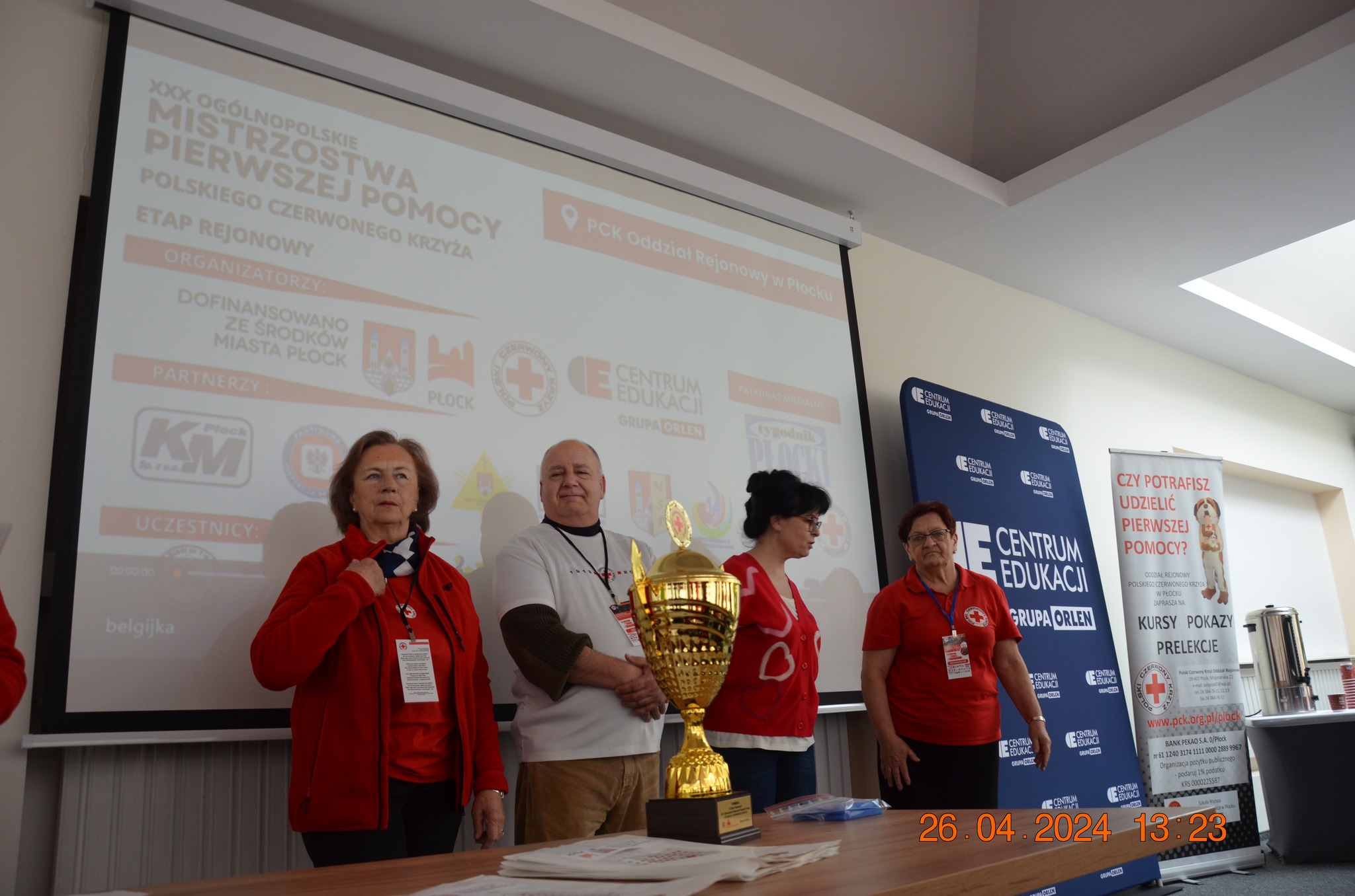 Grupa organizatorów i wolontariuszy PCK stoi przy stole z pucharem na tle ekranu i roll-upów podczas etapu rejonowego XXX Ogólnopolskich Mistrzostw Pierwszej Pomocy w Płocku.