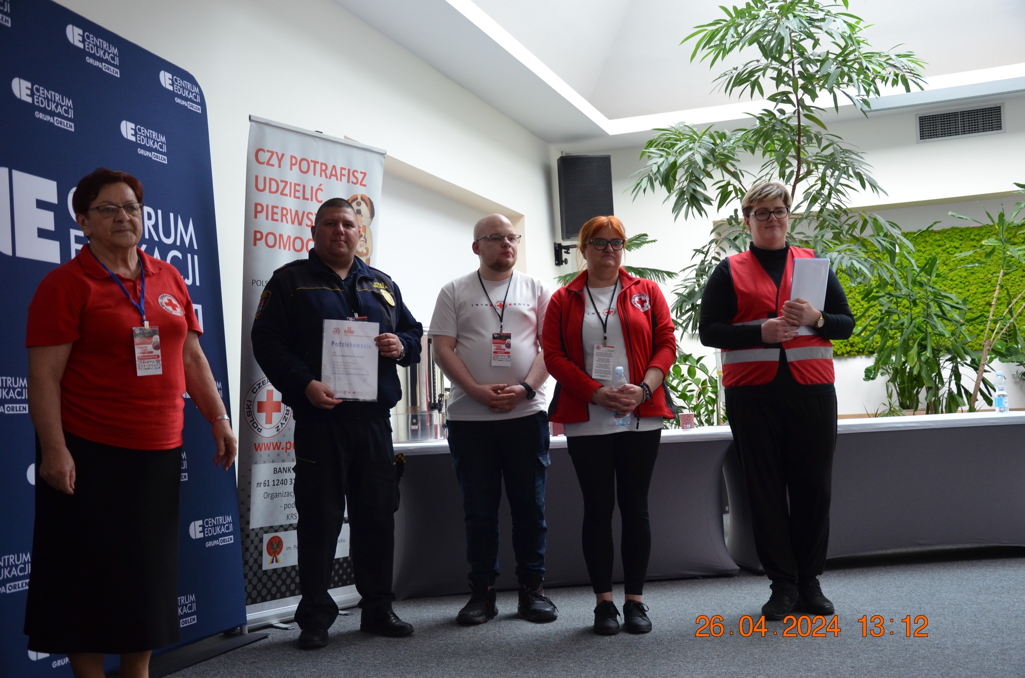 Grupa organizatorów i uczestników etapu rejonowego XXX Ogólnopolskich Mistrzostw Pierwszej Pomocy PCK w Płocku stoi razem podczas oficjalnego podsumowania wydarzenia.