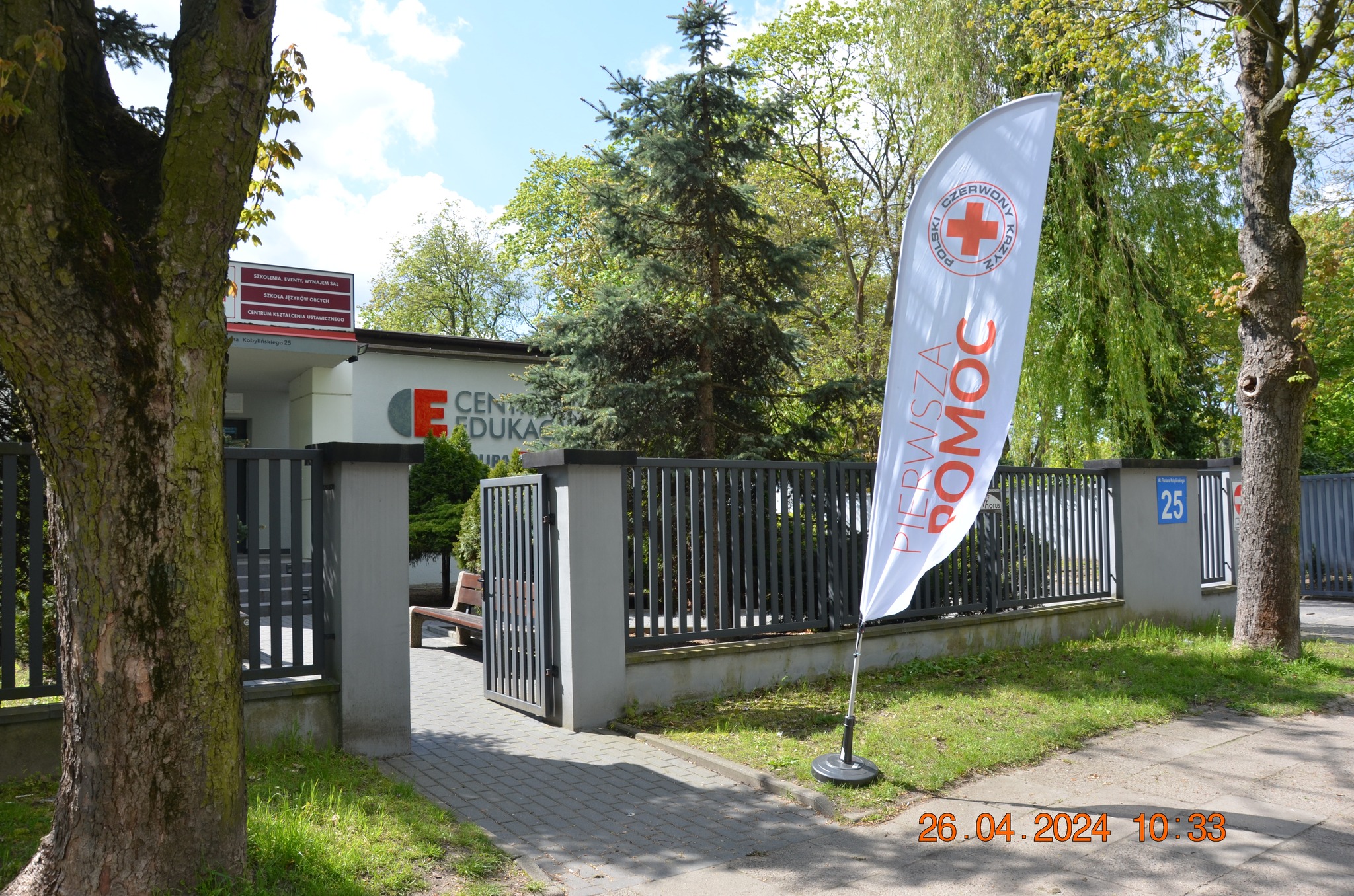 Przed wejściem do Centrum Edukacji Medycznej w Płocku stoi flaga Polskiego Czerwonego Krzyża z napisem „Pierwsza pomoc”, ustawiona podczas rejonowego etapu XXX Ogólnopolskich Mistrzostw Pierwszej Pomocy.