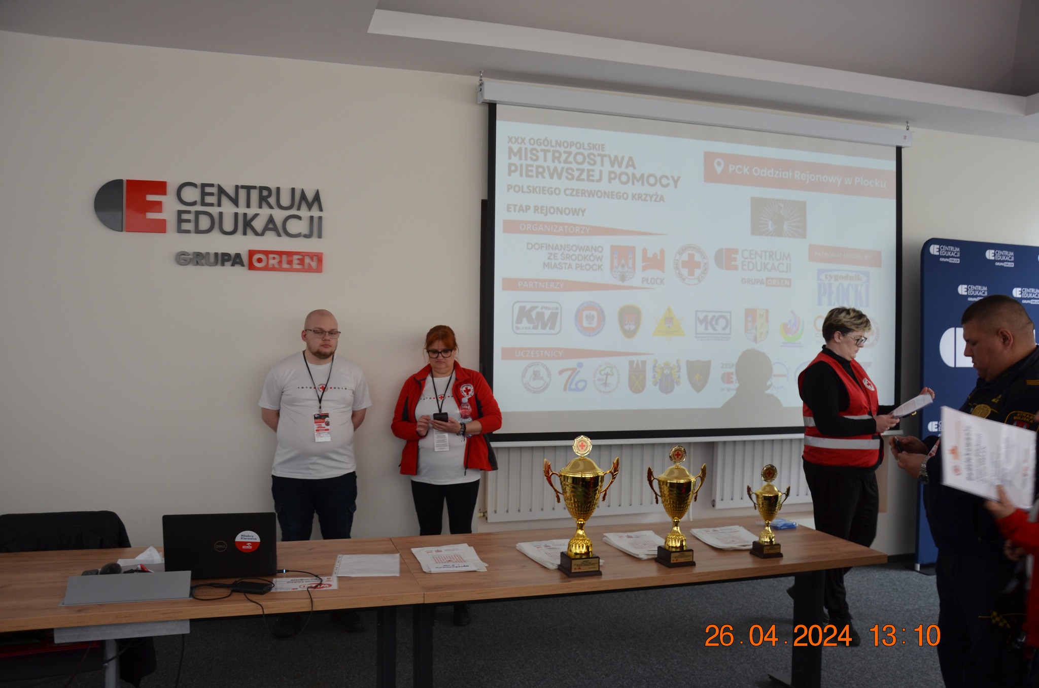 Osoby z Polskiego Czerwonego Krzyża stoją przy stole z pucharami podczas etapu rejonowego XXX Ogólnopolskich Mistrzostw Pierwszej Pomocy w Centrum Edukacji Grupy ORLEN w Płocku.