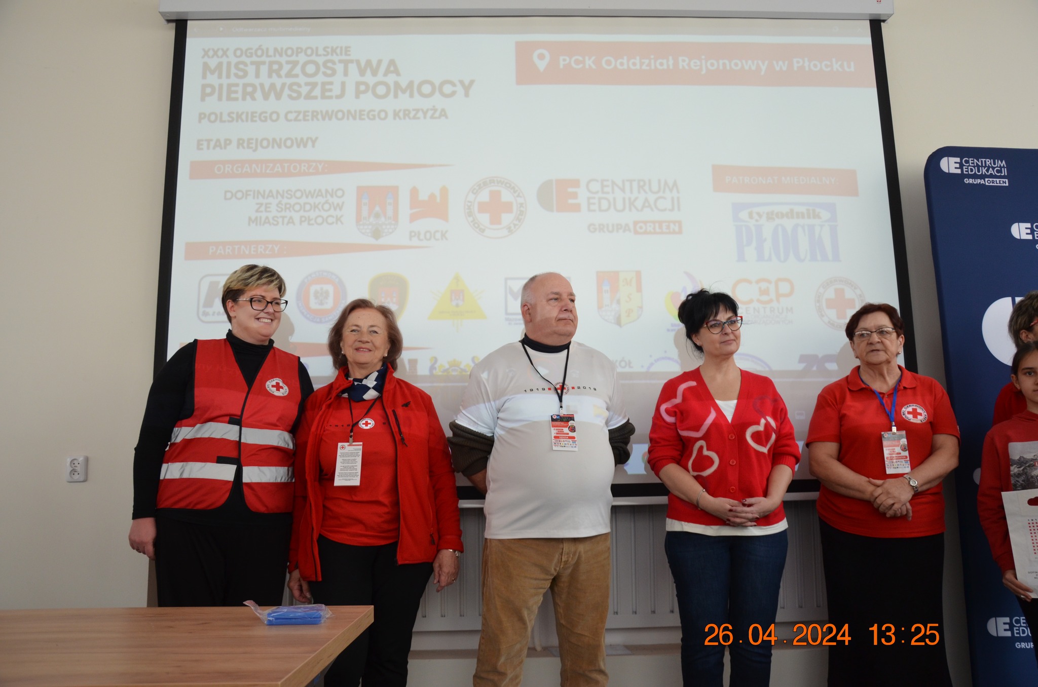 Grupa organizatorów i wolontariuszy PCK stoi przed ekranem z prezentacją podczas etapu rejonowego XXX Ogólnopolskich Mistrzostw Pierwszej Pomocy PCK w Płocku.