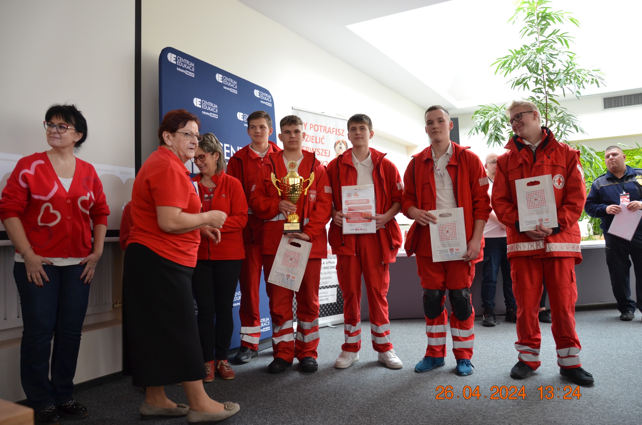 Grupa młodzieży i opiekunów w czerwonych strojach PCK pozuje z pucharem i dyplomami podczas etapu rejonowego XXX Ogólnopolskich Mistrzostw Pierwszej Pomocy PCK w Płocku.