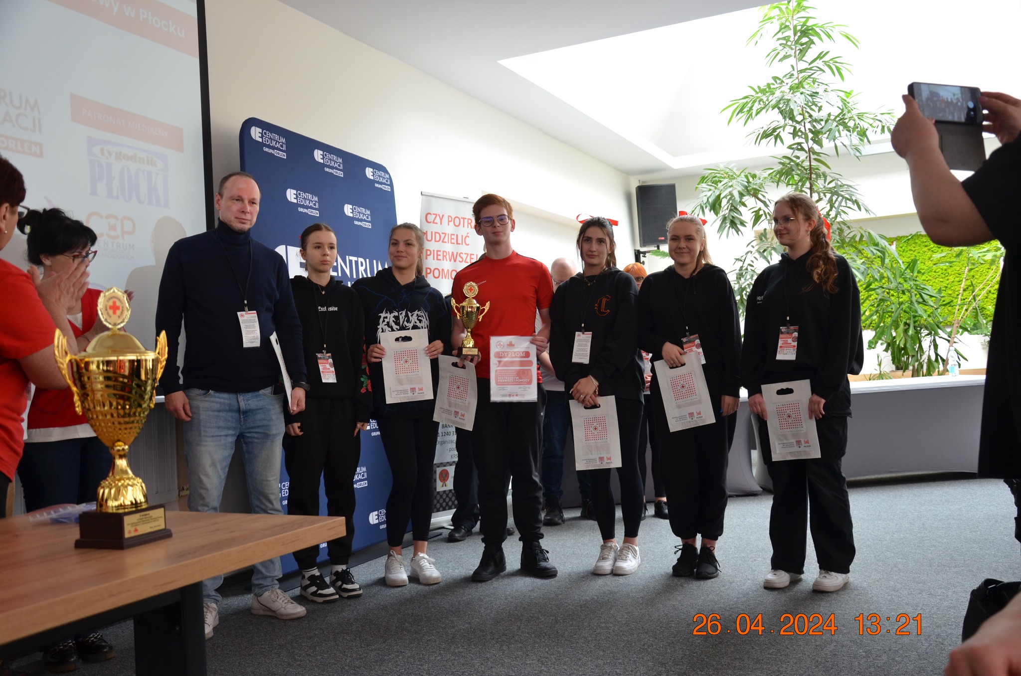 Grupa młodych uczestników z opiekunem stoi na scenie podczas etapu rejonowego XXX Ogólnopolskich Mistrzostw Pierwszej Pomocy PCK w Płocku, trzymając puchary, dyplomy i upominki.