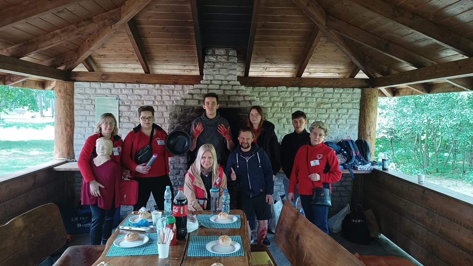 Grupa wicemistrzów pierwszej pomocy Mazowsza pozuje do wspólnego zdjęcia w drewnianej altanie z fantomem i sprzętem medycznym.