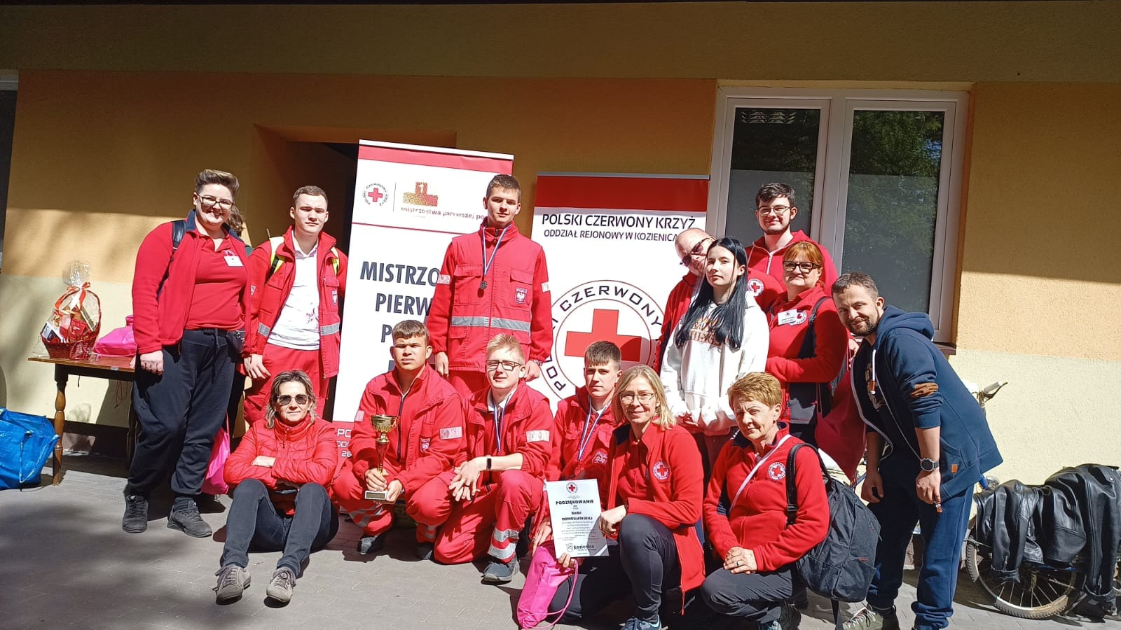 Grupa młodych wolontariuszy PCK w czerwonych strojach pozująca z pucharem i dyplomem przed banerami „Mistrzostwa Pierwszej Pomocy” jako wicemistrzowie pierwszej pomocy Mazowsza.
