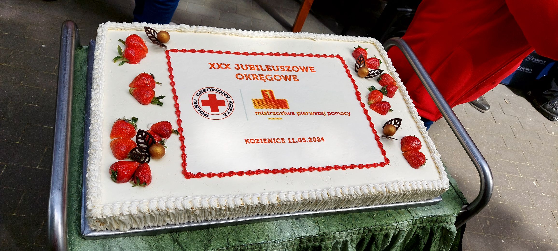 Duży prostokątny tort z truskawkami i napisem „XXX Jubileuszowe Okręgowe Mistrzostwa Pierwszej Pomocy, Kozienice 11.05.2024” przygotowany z okazji wyróżnień wicemistrzów pierwszej pomocy Mazowsza.