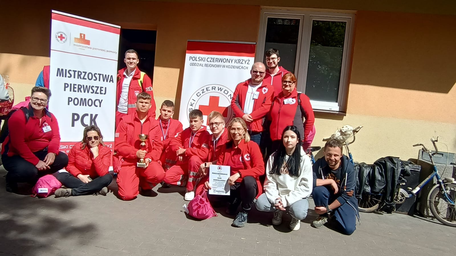 Grupa młodych ratowników PCK pozuje z pucharami i dyplomem na tle banerów „Mistrzostwa Pierwszej Pomocy PCK”, świętując tytuł wicemistrzów pierwszej pomocy Mazowsza.