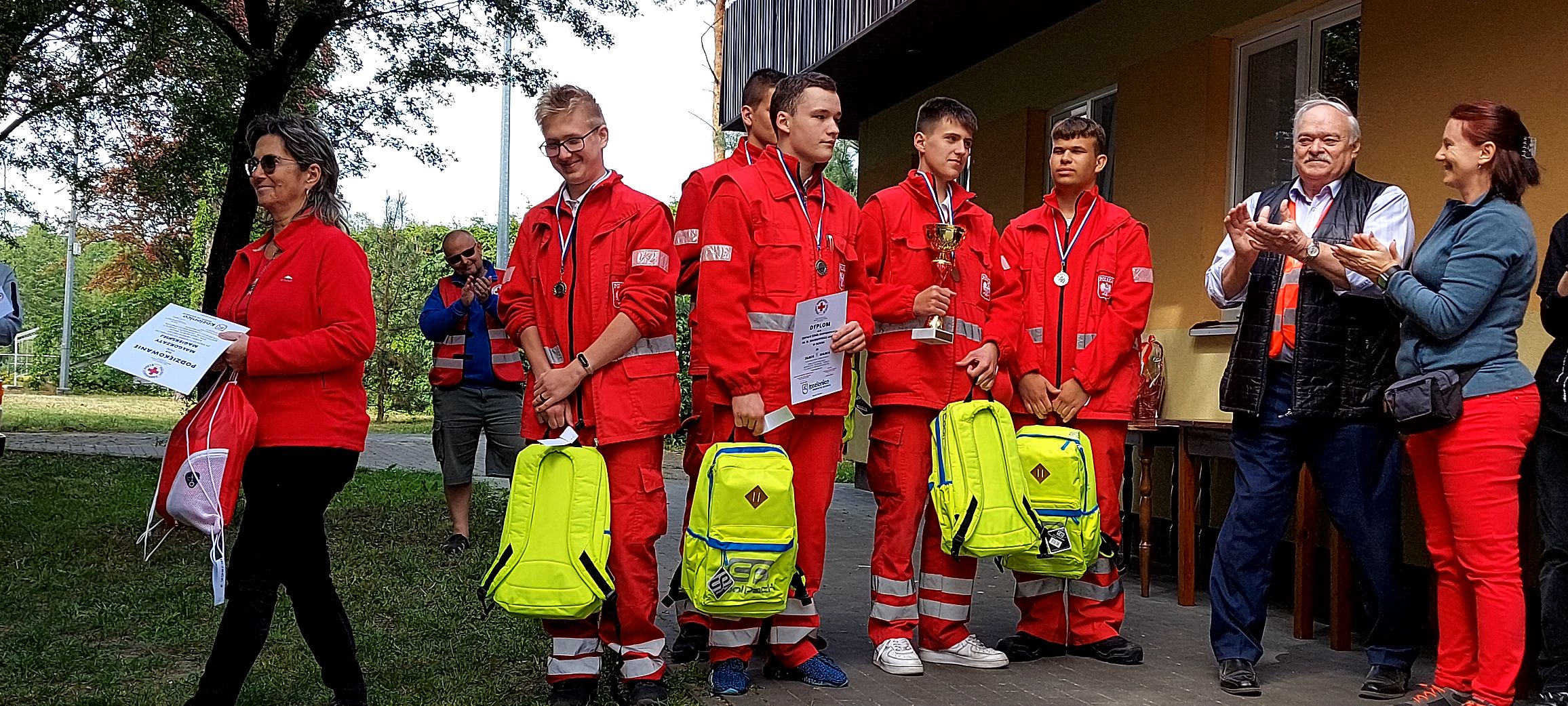 Grupa młodych ratowników w czerwonych mundurach odbiera medale, dyplomy, puchar i żółte plecaki jako wicemistrzowie pierwszej pomocy Mazowsza, stojąc przed budynkiem i oklaskiwana przez opiekunów.