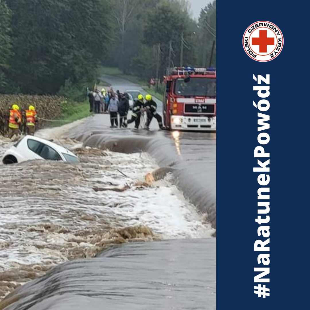 Strażacy ratują ludzi z zalanej przez powódź drogi, obok której nurt niemal całkowicie zatopił samochód; po prawej widoczne jest logo Polskiego Czerwonego Krzyża i napis „#NaRatunekPowódź”.