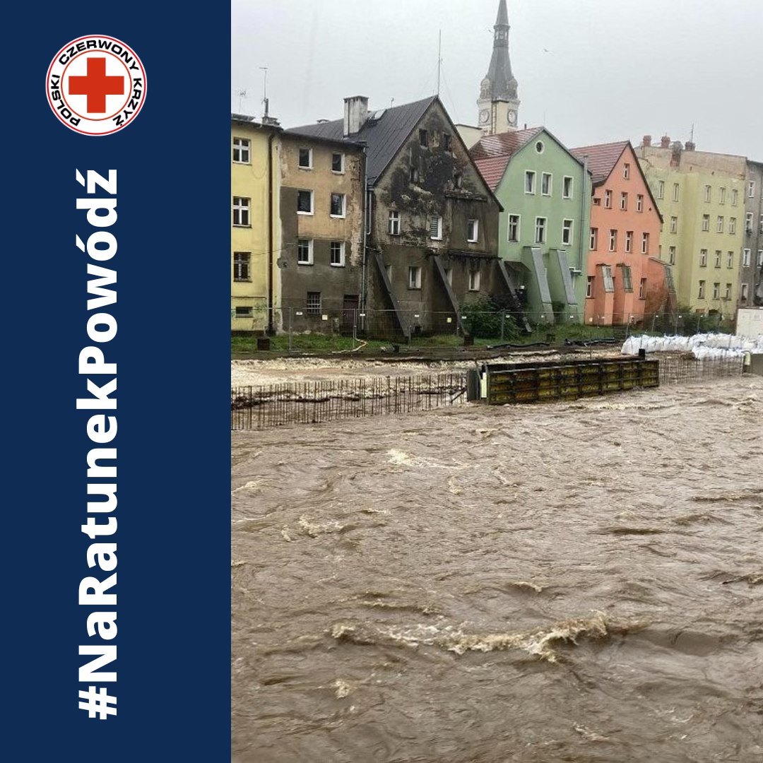 Widok zalanego miasta podczas powodzi z wezbraną rzeką podmywającą kamienice, na grafice z logo Polskiego Czerwonego Krzyża i hashtagiem „#NaRatunekPowódź”.