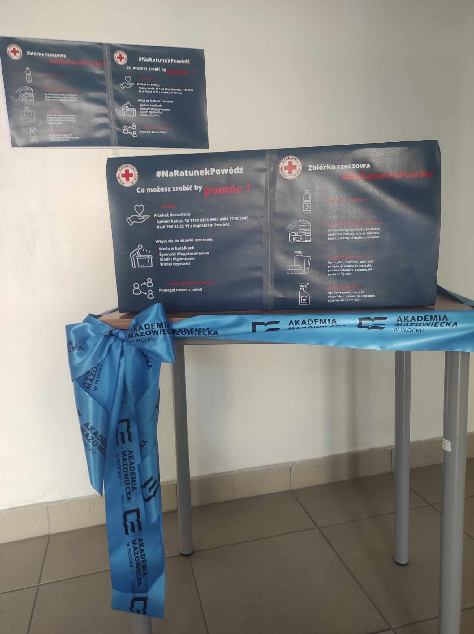 Stolik oznaczony materiałami Akademii Mazowieckiej z dużym plakatem Polskiego Czerwonego Krzyża informującym o zbiórce #NaRatunekPowódź i sposobach pomocy poszkodowanym.