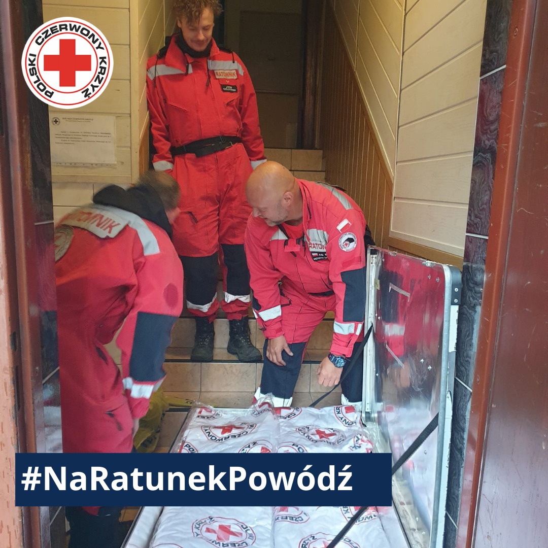 Ratownicy Polskiego Czerwonego Krzyża w czerwonych kombinezonach montują zabezpieczenie przeciwpowodziowe w wejściu do budynku, niosąc pomoc poszkodowanym.