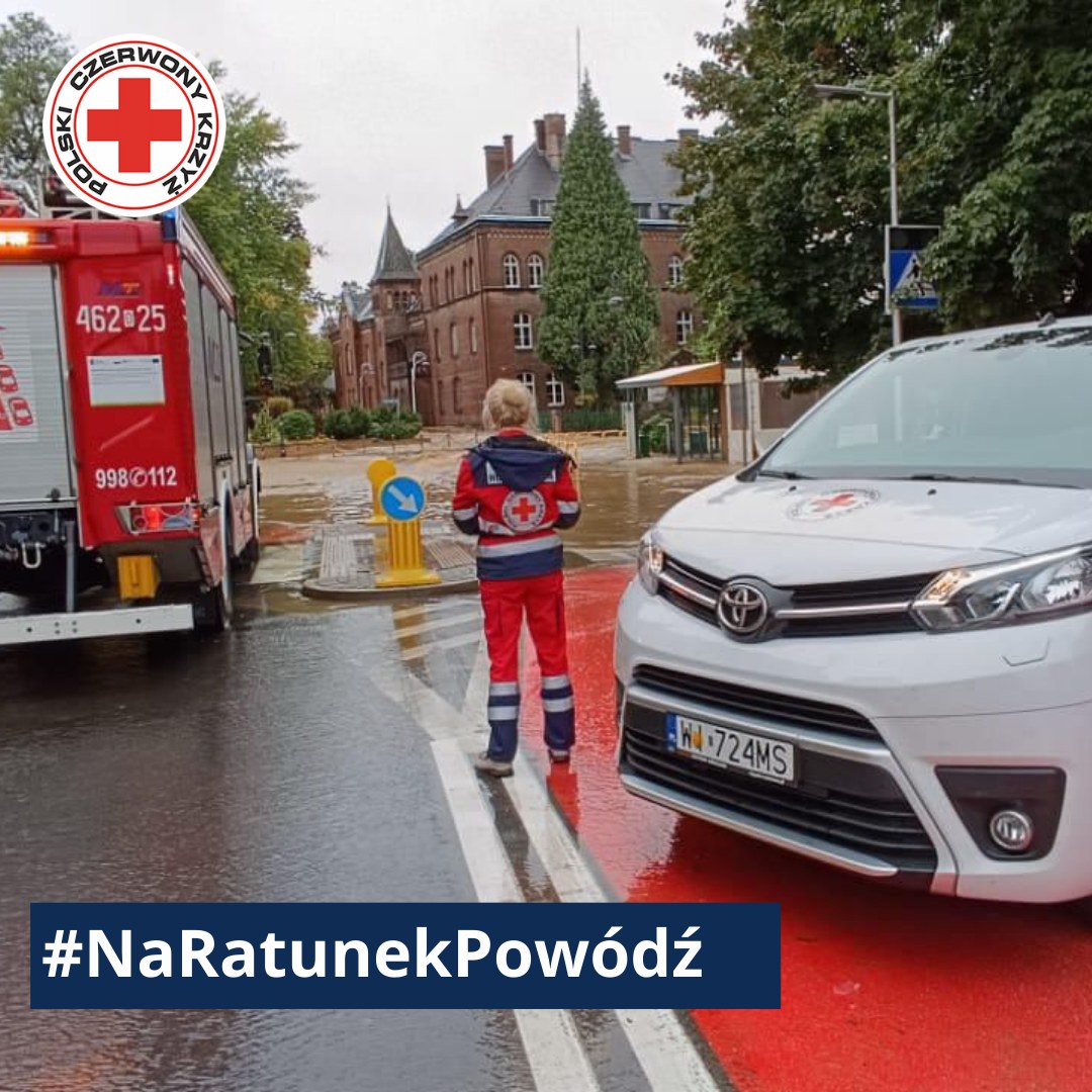 Ratownik Polskiego Czerwonego Krzyża stoi między pojazdem PCK a wozem strażackim na zalanej ulicy, udzielając pomocy podczas powodzi.