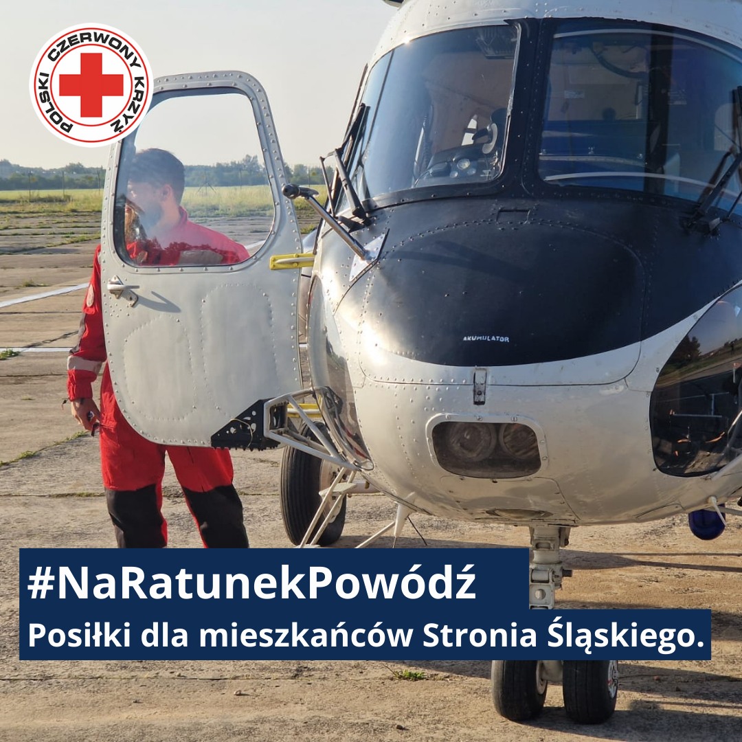 Ratownik Polskiego Czerwonego Krzyża w czerwonym kombinezonie stoi przy śmigłowcu, przygotowując pomoc żywnościową dla mieszkańców zalanego Stronia Śląskiego.