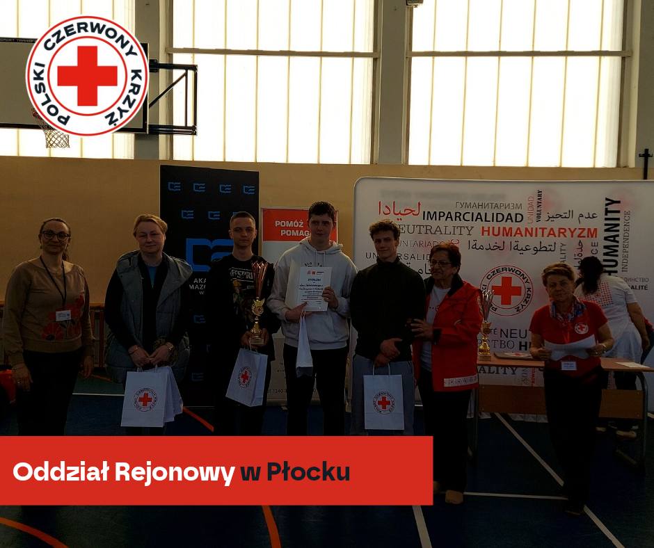 Grupa uczestników i organizatorów XXXII Mistrzostw Pierwszej Pomocy PCK – etap rejonowy w Płocku – pozuje w hali sportowej z pucharami i dyplomami na tle baneru Polskiego Czerwonego Krzyża.