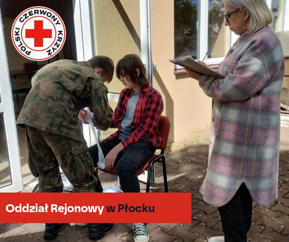 Instruktor w mundurze wojskowym opatruje rękę siedzącej osoby podczas ćwiczeń pierwszej pomocy PCK w Płocku, a obok stoi sędzia z notatnikiem.