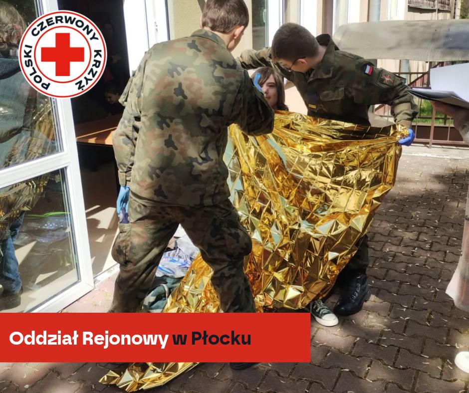 Uczestnicy XXXII Mistrzostw Pierwszej Pomocy PCK w Płocku w wojskowych mundurach okrywają poszkodowaną złotym kocem termicznym podczas pozoracji medycznej.