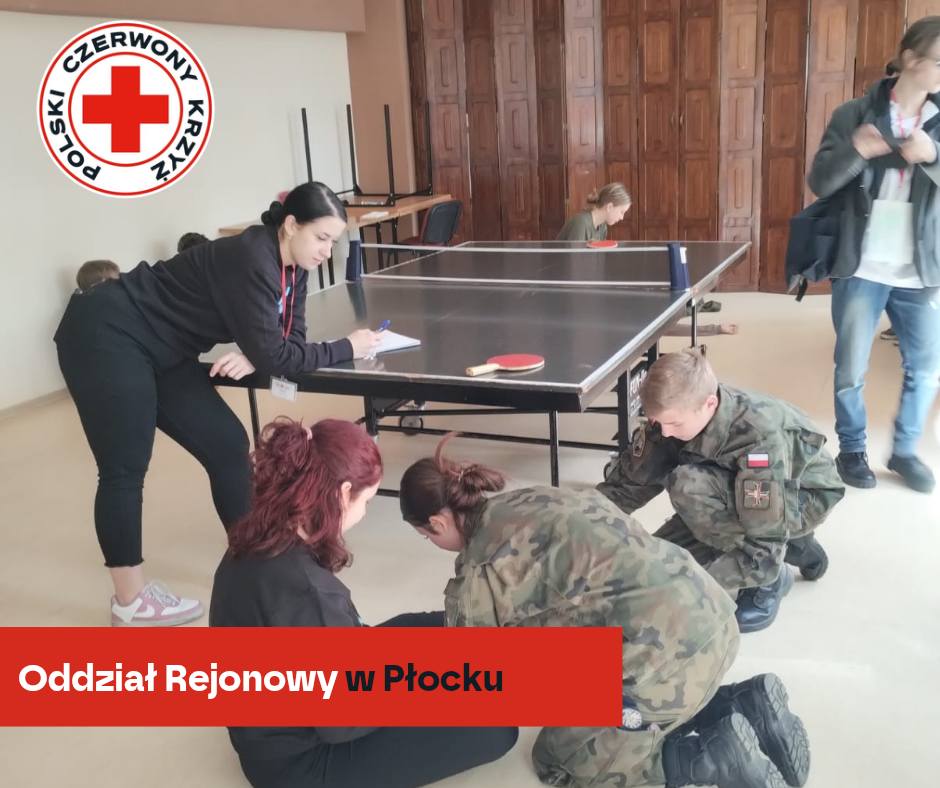 Uczestnicy XXXII Mistrzostw Pierwszej Pomocy PCK w Płocku trenują udzielanie pierwszej pomocy pod okiem instruktorki w sali z pingpongowym stołem.
