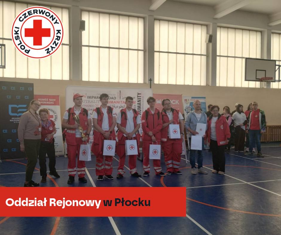 Grupa uczestników i organizatorów XXXII Rejonowych Mistrzostw Pierwszej Pomocy PCK w Płocku pozuje w hali sportowej z dyplomami i torbami z logo Polskiego Czerwonego Krzyża.