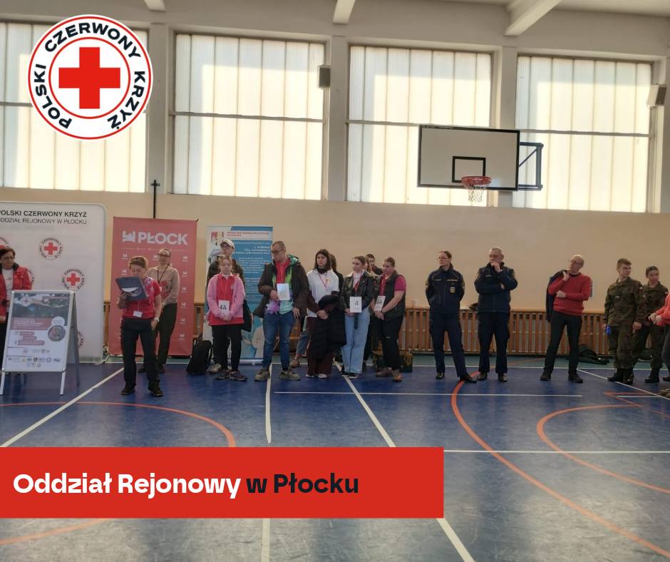 Uczestnicy XXXII Rejonowych Mistrzostw Pierwszej Pomocy PCK w Płocku stoją w rzędzie w hali sportowej podczas oficjalnej części wydarzenia.