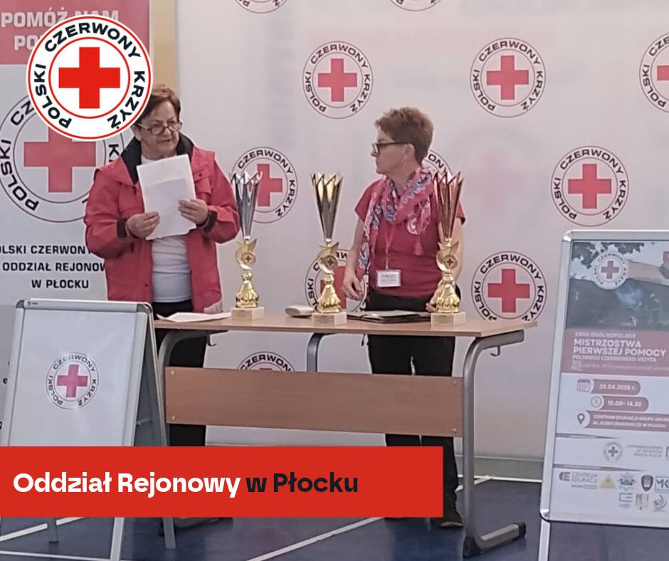 Dwie przedstawicielki Polskiego Czerwonego Krzyża stoją przy stole z pucharami podczas etapu rejonowego XXXII Mistrzostw Pierwszej Pomocy PCK w Płocku.