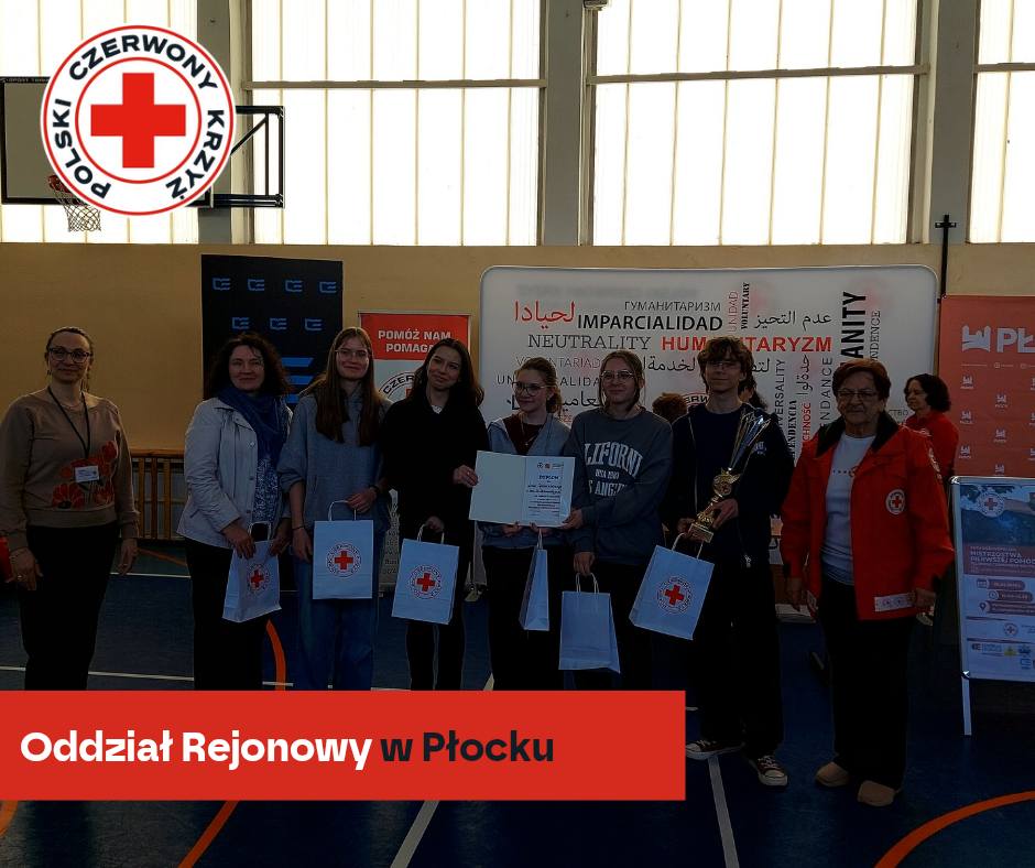 Grupa uczestników XXXII Mistrzostw Pierwszej Pomocy PCK w Płocku pozuje w sali gimnastycznej z dyplomem, pucharami i torbami z logo Polskiego Czerwonego Krzyża.