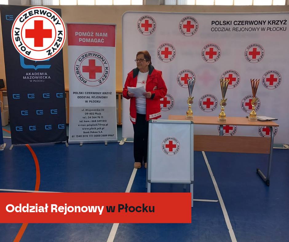 Kobieta w czerwonej kurtce PCK stoi z kartkami przy stoliku z pucharami na tle banerów Polskiego Czerwonego Krzyża podczas XXXII Mistrzostw Pierwszej Pomocy, etap rejonowy w Płocku 2026.