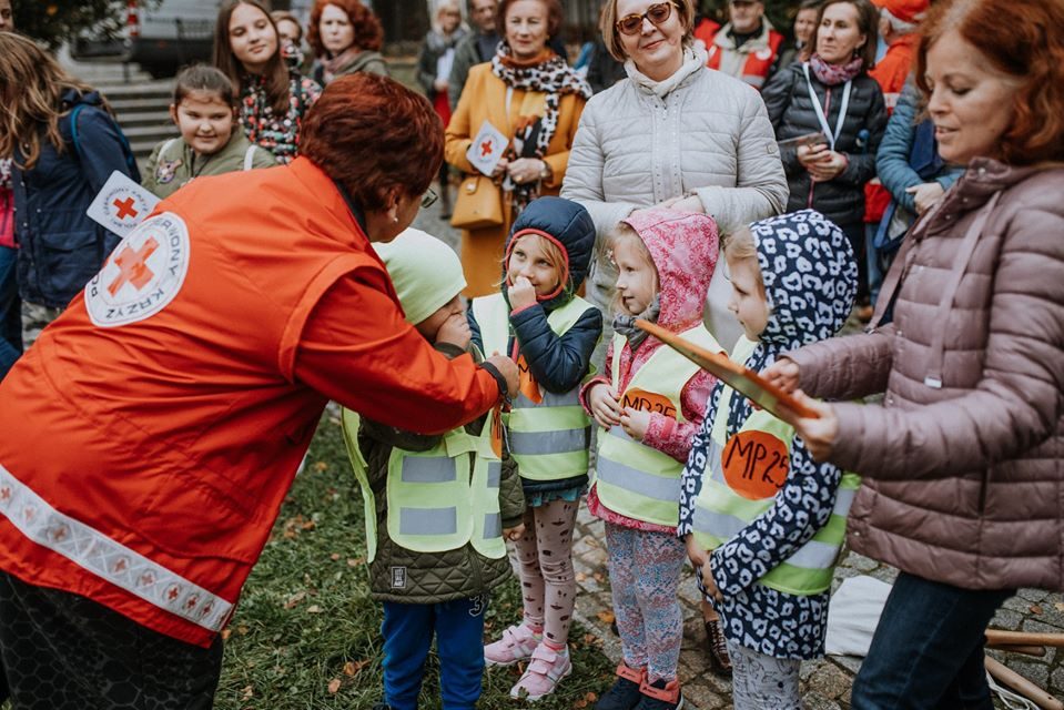 Wolontariuszka Polskiego Czerwonego Krzyża wręcza nagrody uśmiechniętym przedszkolakom w odblaskowych kamizelkach podczas plenerowego wydarzenia z udziałem mieszkańców.