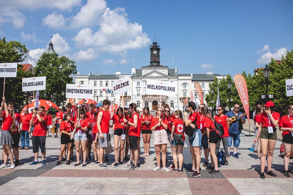 Grupa młodych wolontariuszy PCK w czerwonych koszulkach stoi na rynku przed ratuszem, trzymając tabliczki z nazwami województw.