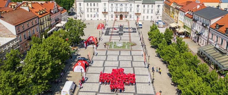 Widok z lotu ptaka na rynek miasta z ratuszem, gdzie grupa osób w czerwonych koszulkach tworzy na placu duży krzyż symbolizujący Polski Czerwony Krzyż.