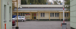 Wejście do siedziby PCK w jednokondygnacyjnym budynku z zadaszonym podjazdem i parkingiem przed głównymi drzwiami.